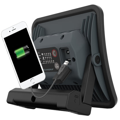 Product photo of a dark gray SCANGRIP work light on an isolated white background powering a white smartphone displaying a green charging battery icon, with visible USB port and control panel showing brightness percentages and CCT Kelvin settings