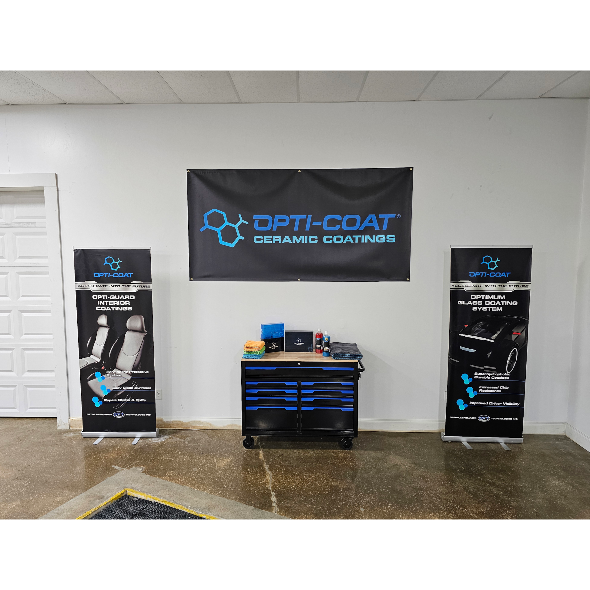 Photograph of an Opti-Coat ceramic coatings display in a white garage showroom featuring a large black banner with blue text two matching pull-up banners for interior and glass coatings and a black and blue toolbox with colorful towels and product bottles