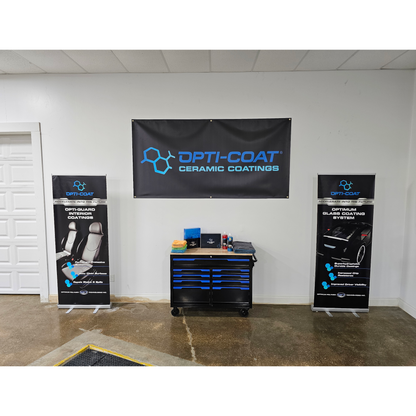 Photograph of an Opti-Coat ceramic coatings display in a white garage showroom featuring a large black banner with blue text two matching pull-up banners for interior and glass coatings and a black and blue toolbox with colorful towels and product bottles