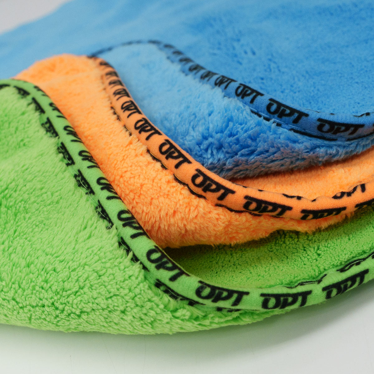 Close-up photograph of three vibrantly colored stacked microfiber towels in green, orange, and blue, each featuring a black trim with the repeating OPT logo, showcasing their soft plush texture