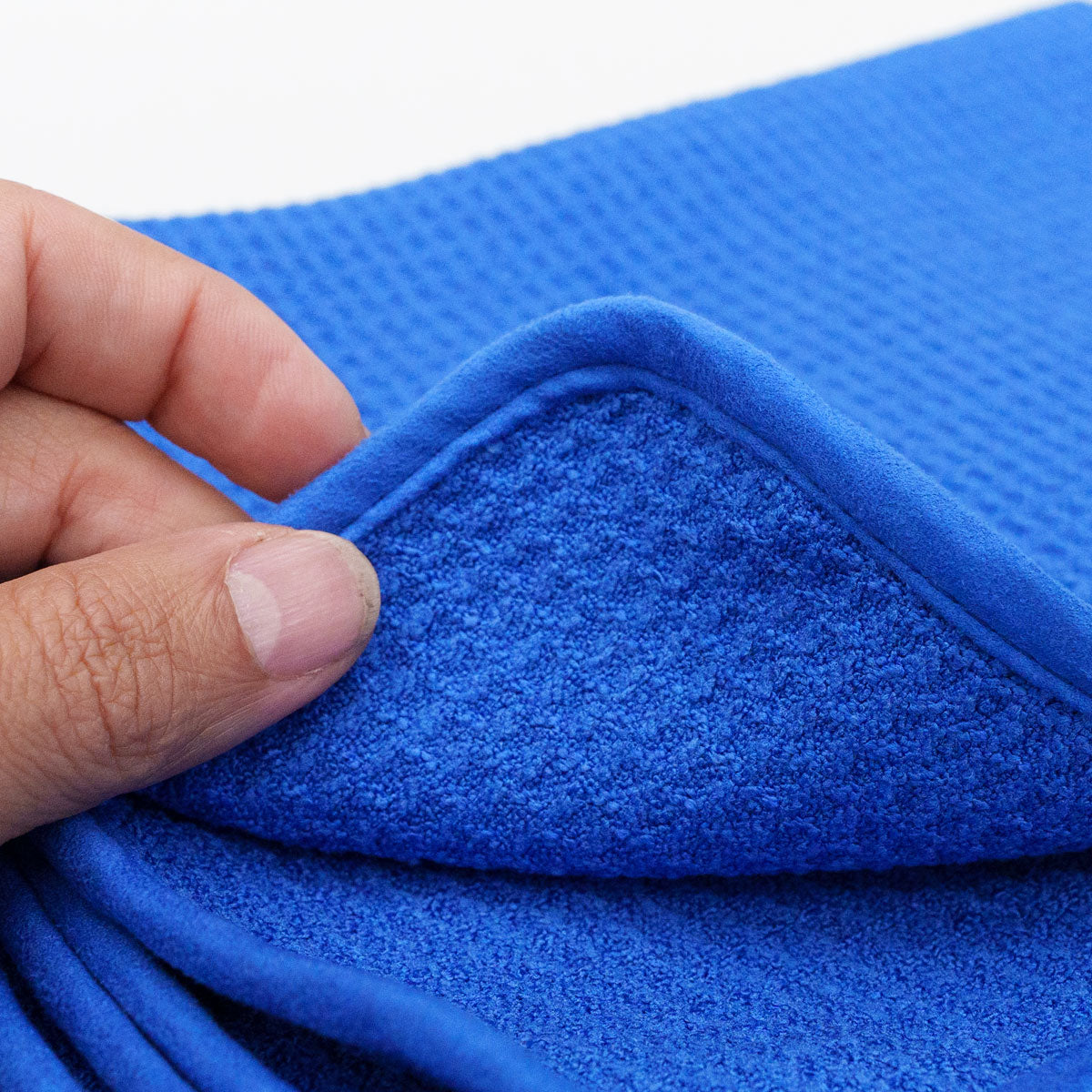 Photograph of a hand holding a corner of a bright royal blue microfiber towel, revealing a textured waffle weave on one side and a soft, looped pile on the other with a neatly stitched edge