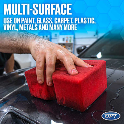 A hand uses two red multi-surface applicator sponges to clean a wet dark gray car hood with water droplets, featuring text overlay "MULTI-SURFACE USE ON PAINT, GLASS, CARPET, PLASTIC, VINYL, METALS AND MANY MORE" and an OPT logo