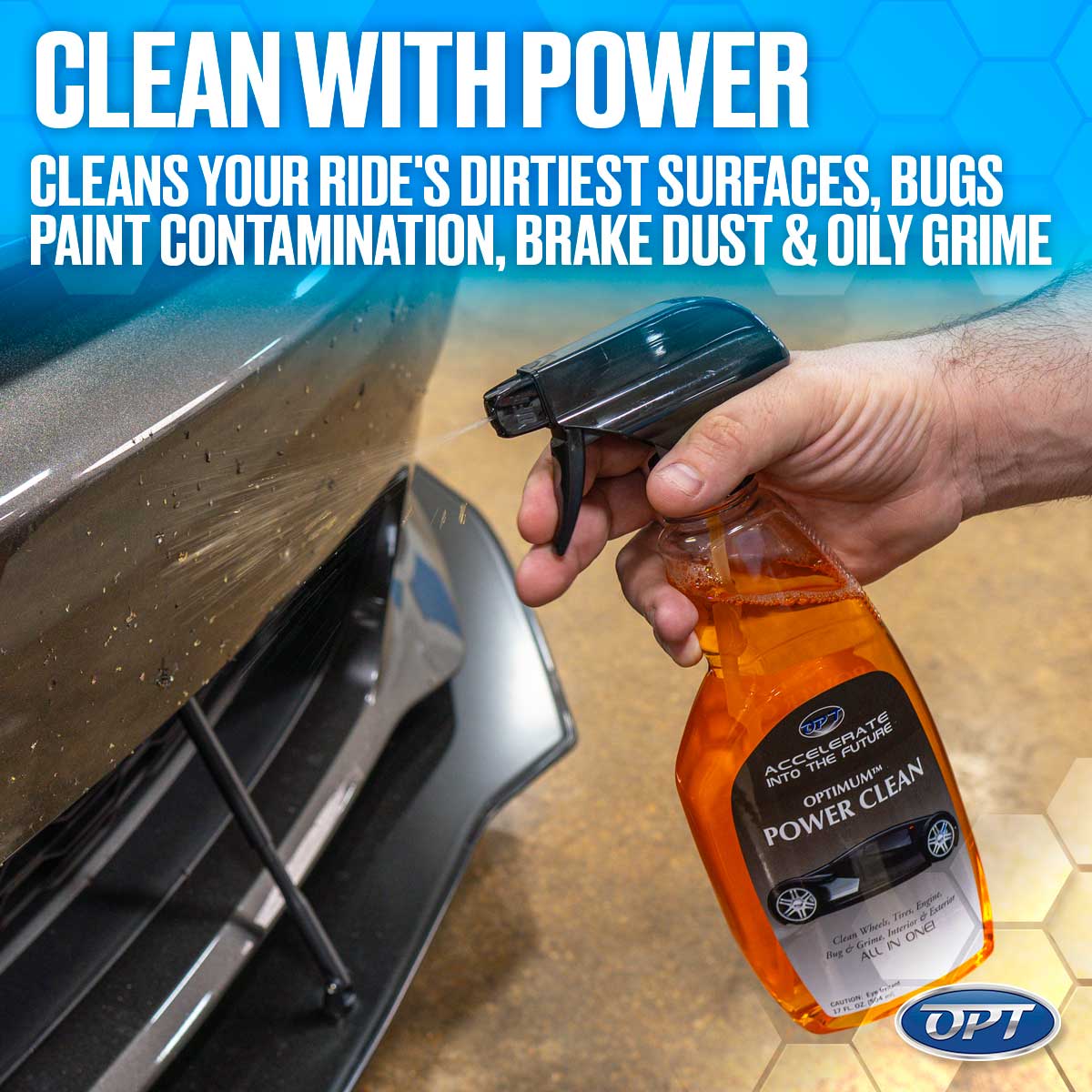 A hand sprays orange Optimum Power Clean from a clear bottle onto a dirty grey car bumper, actively cleaning visible grime and bugs off the vehicle with a powerful solution