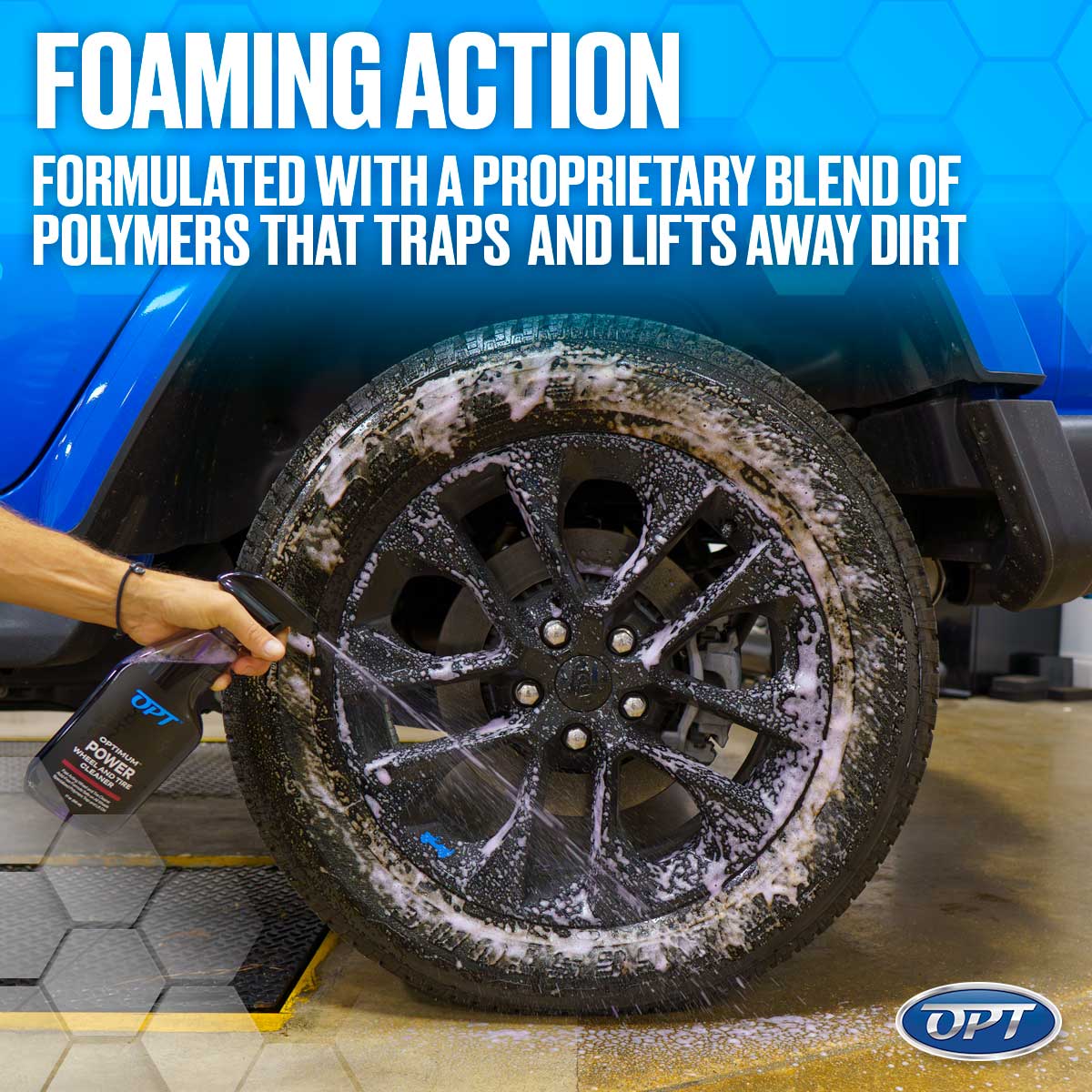A person's hand sprays OPTimum Power Wheel and Tire Cleaner from a black bottle onto a dirty black wheel and tire of a blue Jeep Wrangler, creating white foam, positioned on a yellow and gray hexagonal floor mat
