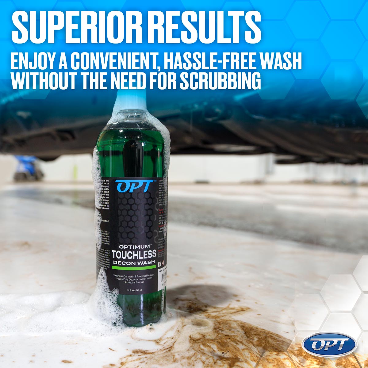 Green Optimum Touchless Decon Wash bottle with overflowing white foam and brown sludge on a tiled garage floor under a dark vehicle, featuring a blue "SUPERIOR RESULTS" banner