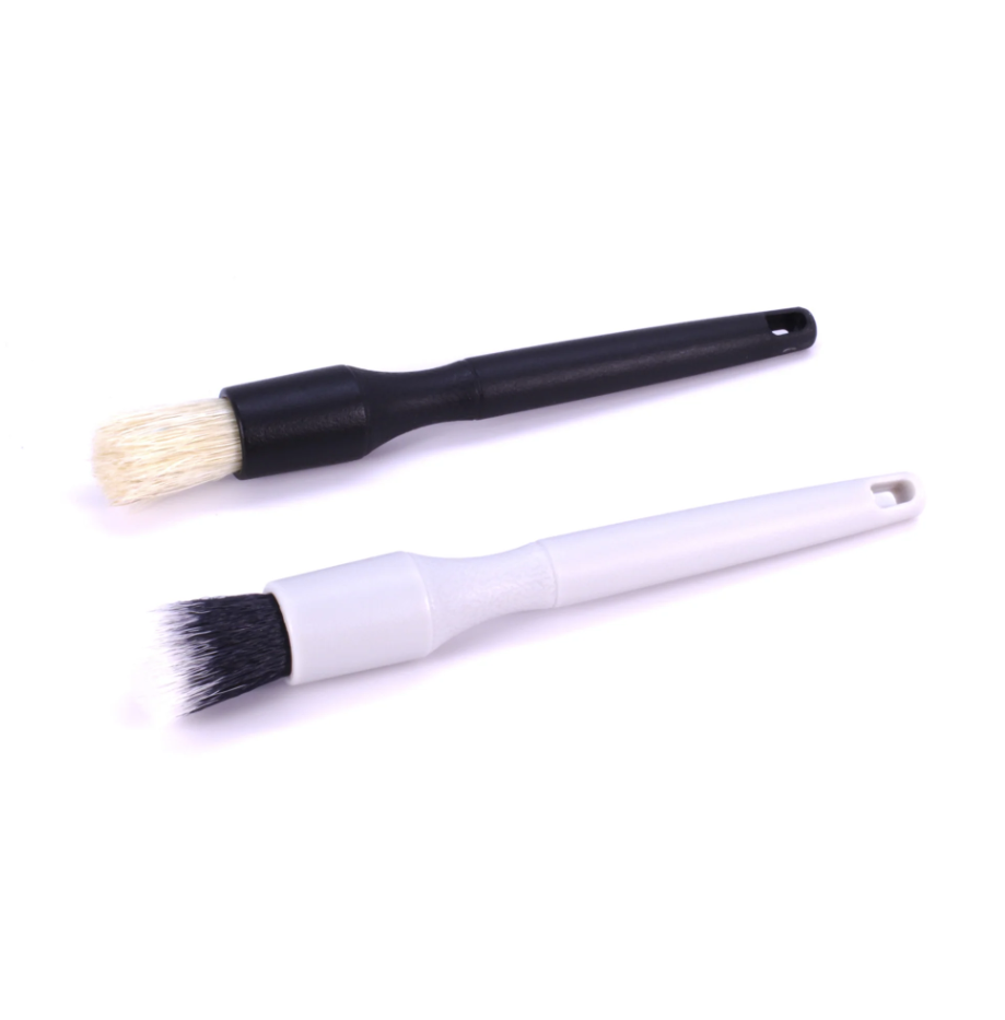 Photography of two detailing brushes on a white background one with a black handle and light beige bristles the other with a white handle and black and white tipped bristles both featuring small hanging holes